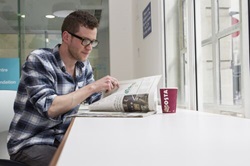 A man sits at a table next to a window. He is drinking a coffee and reading a magazine