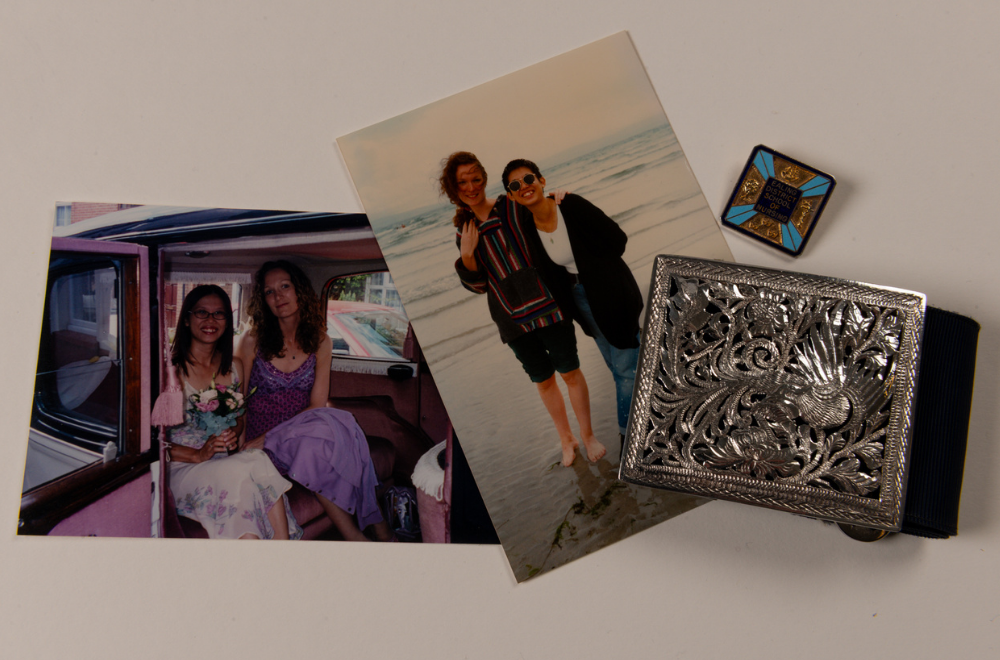Two photos of two women, Maria Tighe with brown curly hair and Tzah Shian with straight black hair - in the left image they are seated in a vehicle and in the right they are standing on a beach. Next to the pictures is a blue nurses belt with a decorative, silver-coloured buckle, as well as a square-shaped gold, dark blue and light blue nurses badge for Ealing District School of Nursing.
