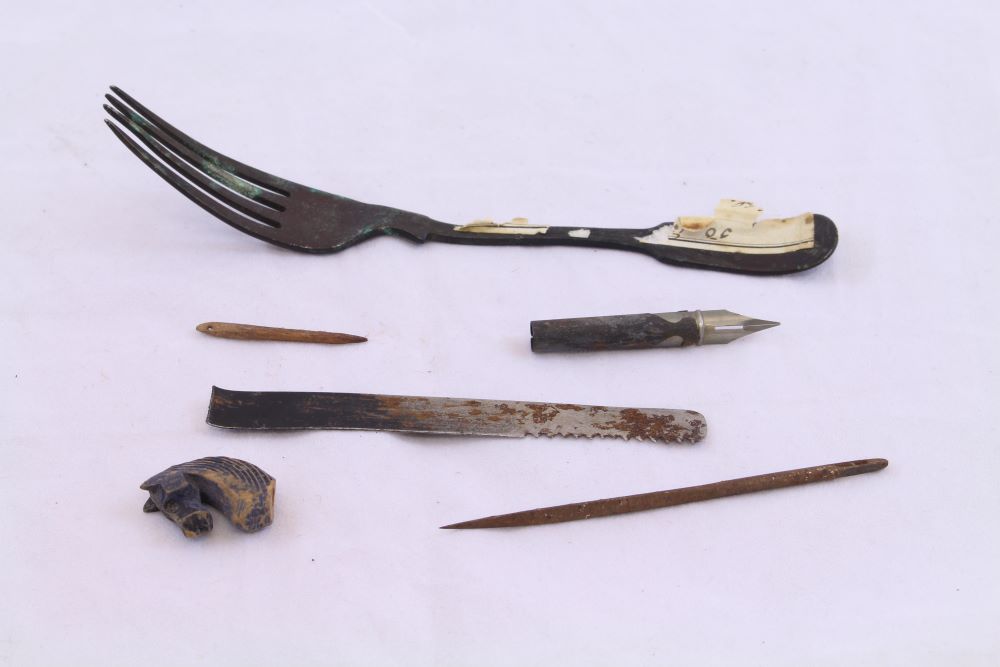 A collection of items swallowed by prisoners. From top to bottom: a fork, a fountain pen nib, a wooden needle, the blade of a knife, a small wooden horses head, a long thin sharp object. 