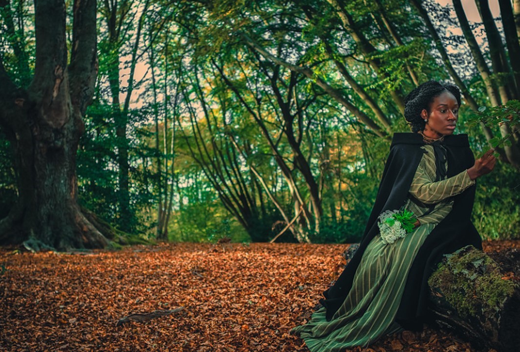 Photograph of a Black woman dressed in a cape and green dress with white stripes, sitting on a tree trunk in the middle of a forest.