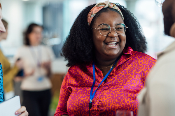 A woman in a vibrant pink and orange patterned blouse, wearing glasses and a headband, smiles while engaged in conversation at a social or networking event.