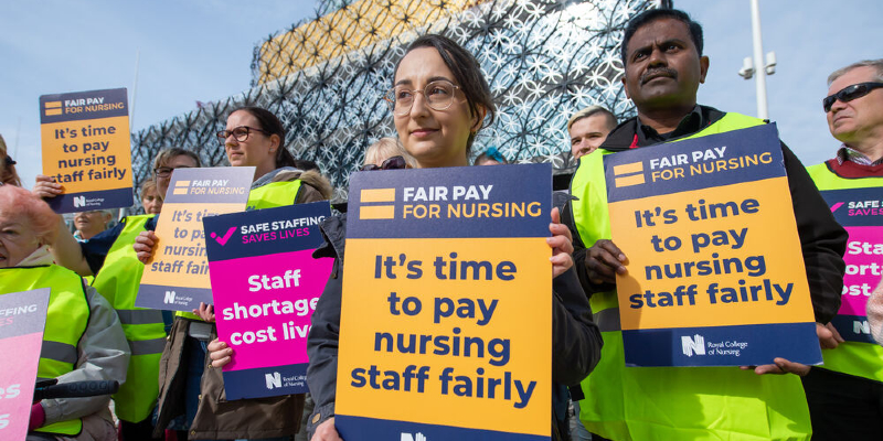 RCN members are seen demonstrating outside over fair pay for nursing. They are holding placards that read "It's time to pay nursing staff fairly" and "Staff shortages cost lives".