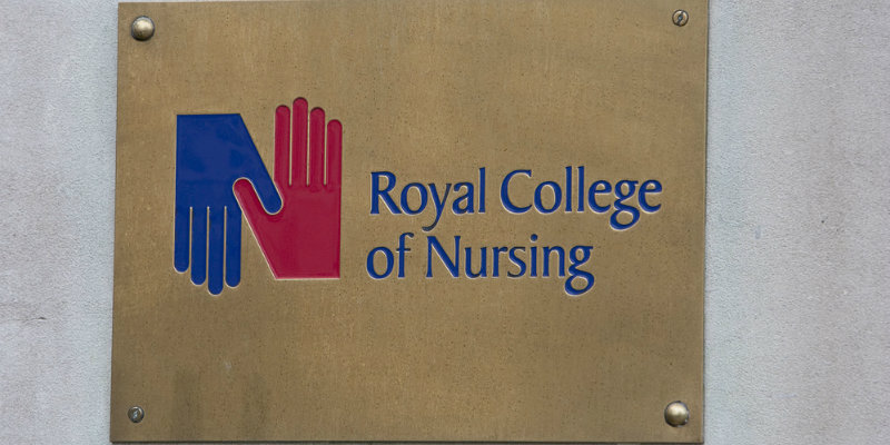 A brass plaque is seen square on, on the outside of a building, with the name "Royal College of Nursing" in blue writing on the right and the RCN logo to the left, a blue hand facing down and a red hand facing up, with their thumbs interlinking