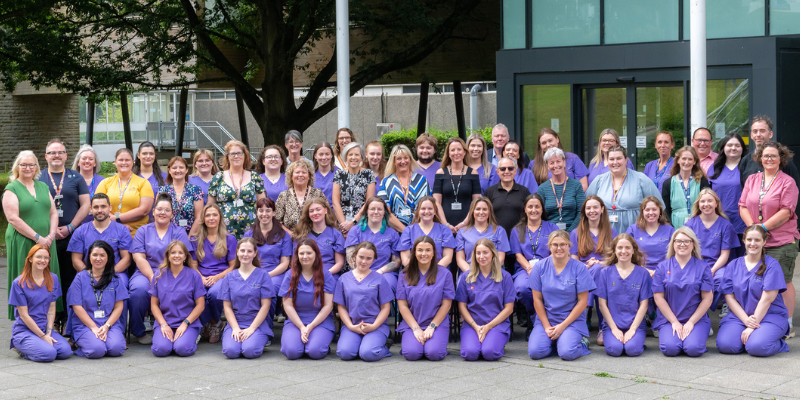 Posed group shot of nursing students and university staff outside university building, all looking toward camera with hands clasped in front of them