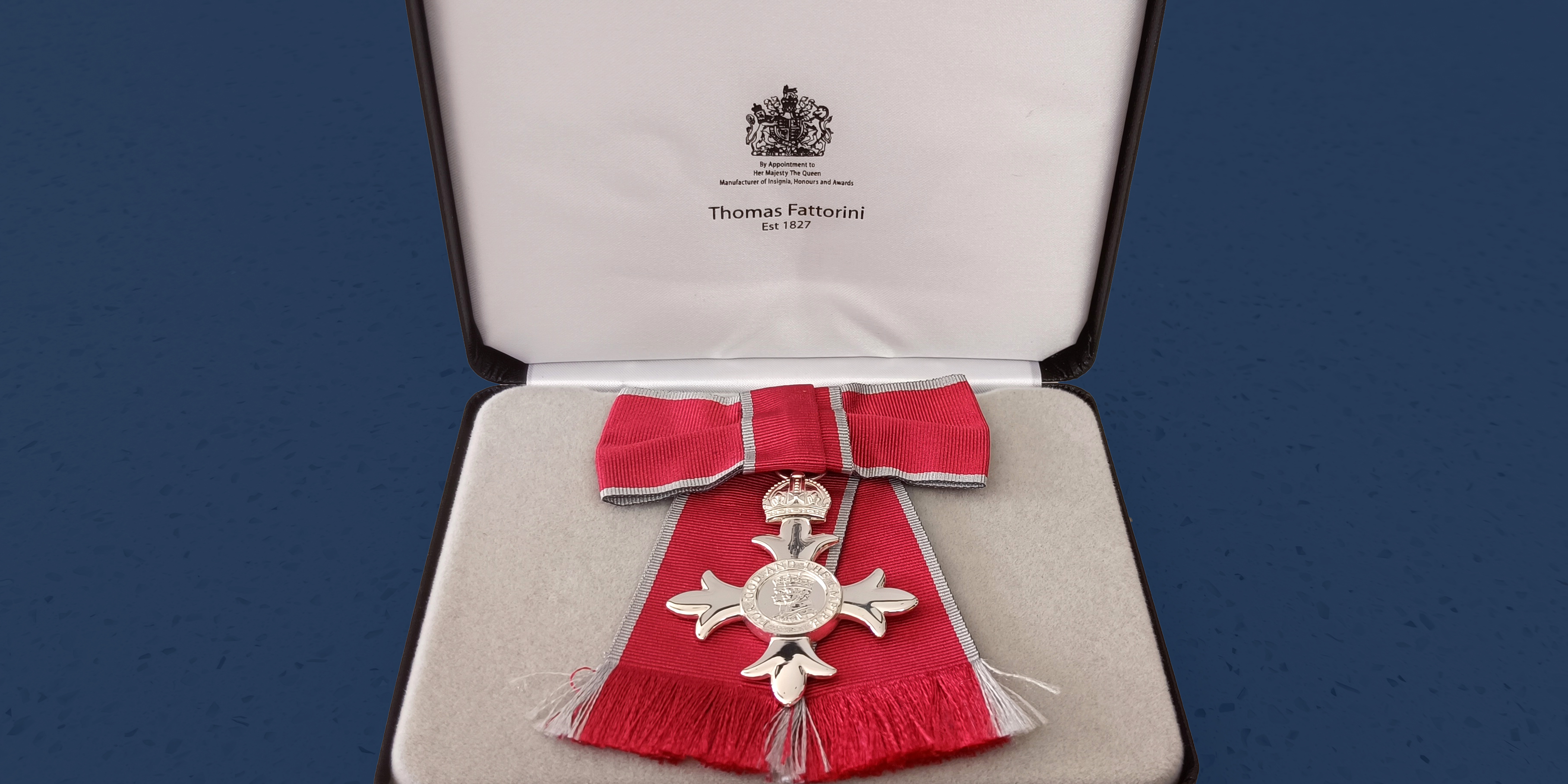 A female MBE medal is shown in its display case. There is an ornate silver cross surrounded by red ribbon, sitting in a plush grey box with an emblem inside the lid. A blue background is visible behind the box.