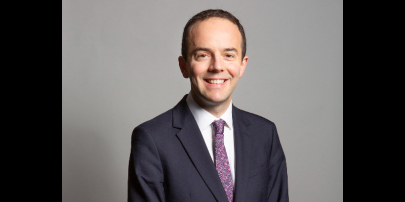 James Murray, the health secretary, is shown in his official House of Commons portrait. He is wearing a dark grey suit, a white shirt and a purple, patterned tie. He looks directly at the camera smiling.