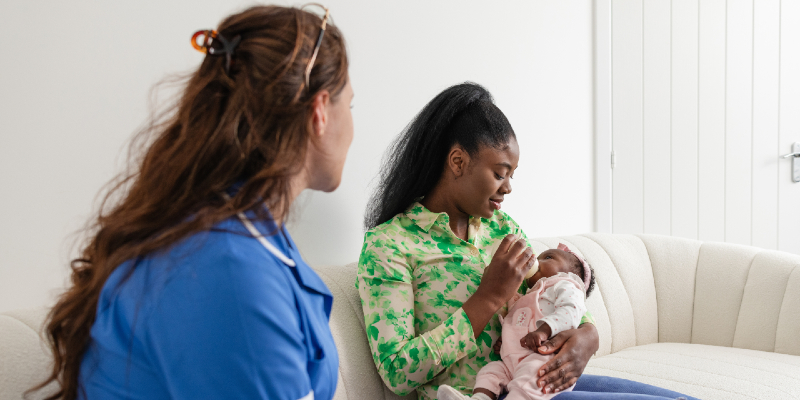 Young female health visitor sitting in a living room with a new mother and her baby girl who is being bottle fed