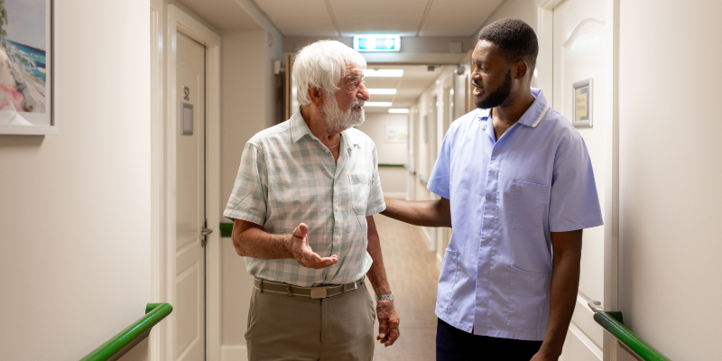 two people walking along a corridor in a residential care home, a elderly resident and a health care worker talking as they walk together