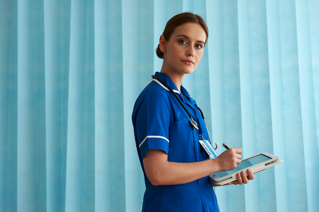 A health care worker stands looking into the camera. She is wearing a blue uniform with short sleeves and behind her is a light blue curtain. She holds a computer tablet and pen or stylus which she is holding to the screen. She has dark brown hair which is tied up.