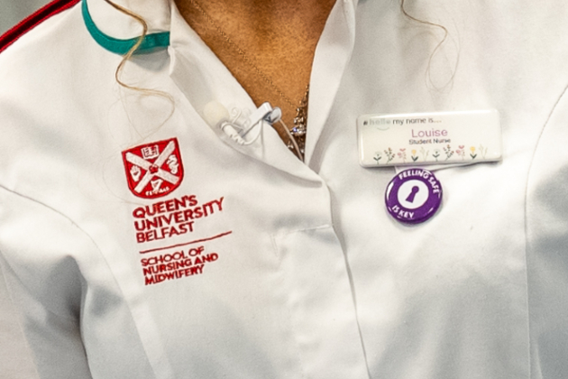 A close up shows a nursing student's white uniform with a purple and white Unlocking Safety badge pinned to her chest. The badge reads: "Feeling safe is key" with a keyhole in the middle. The words "Queen's University Belfast School of Nursing and Midwifery" are embroidered in red, along with the university's crest. There is also a nametag with the name "Louise"