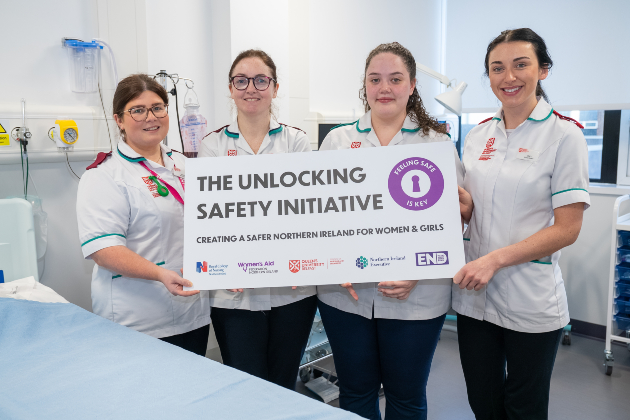 A group of four female nursing students hold a sign reading "Unlocking Safety Initiative". They are all wearing white uniform tops with black trousers. They stand in a hospital setting with a bed and medical equipment visible around them.
