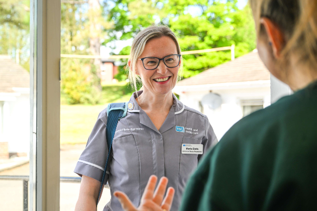 ANP in district nursing Maria Betts is greeted by a patient at the door of their home. She wears a grey uniform and dark-rimmed glasses with her dark blonder hair tied up. She is smiling at the patient