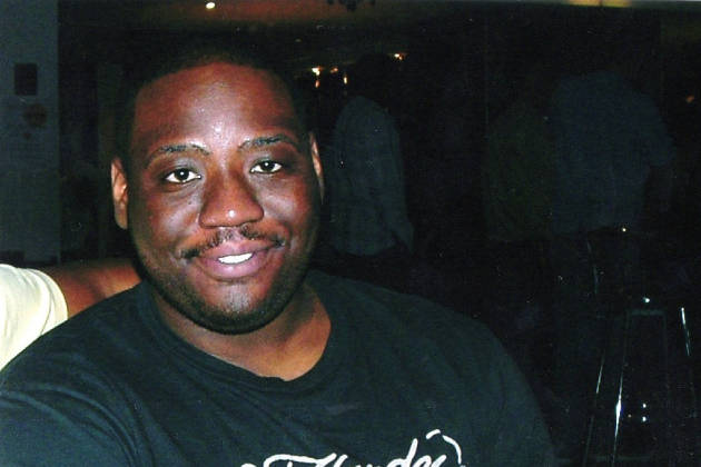 A young man is seen smiling and looking into the camera. He is wearing a black t-shirt