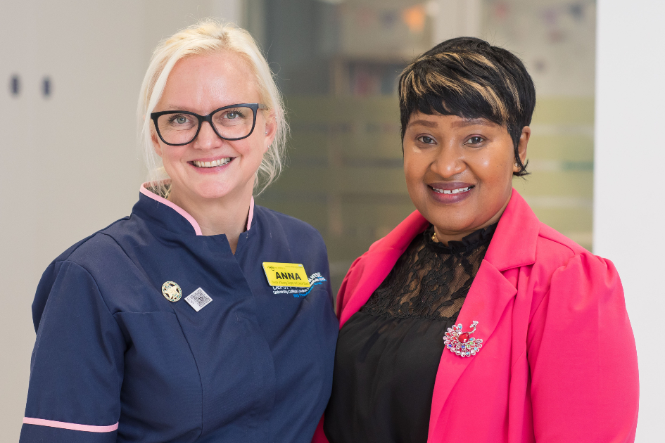 Two nurses stand next to each other facing the camera. Anna, on the left, wears her navy nurse's uniform while Nima, on the right wears her own clothes, a bright pink jacket and black clothes. They are good friends, and are standing close together while smiling.