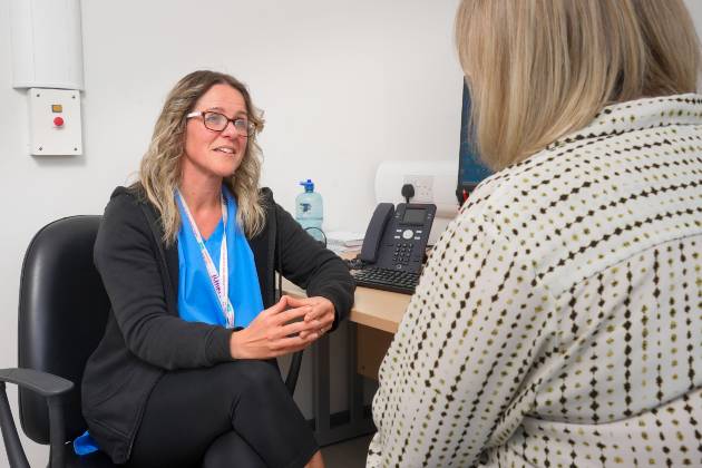 Kelly, a mental health nurse, sits at her desk facing a patient. Kelly is wearing a blue top with a black cardigan over the top. She has glasses and dark blonder hair. She is steepling her fingers and her body language is open as she listens to her patient.