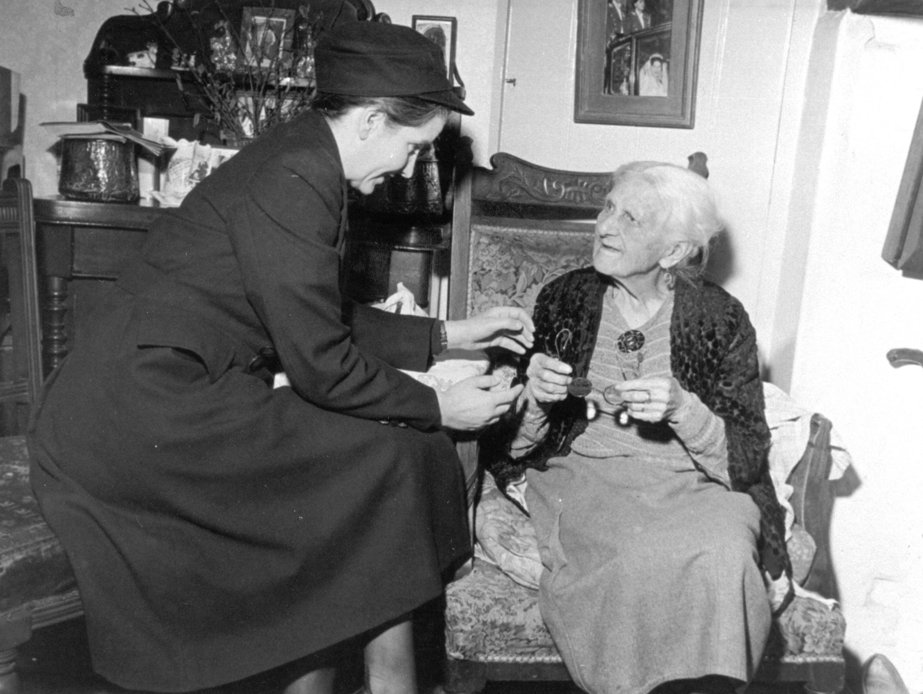 A black and white image from circa 1960s shows a woman district nurse visiting an elderly woman patient in her home