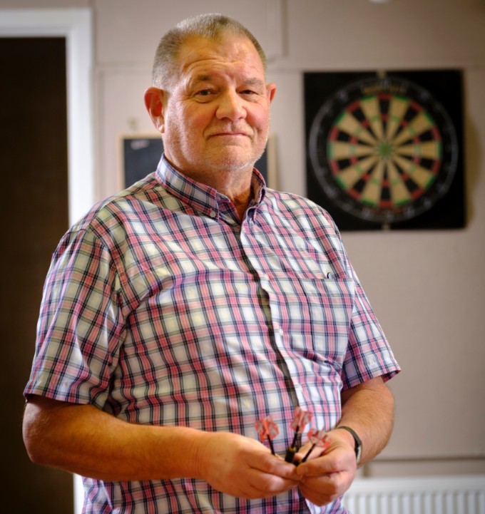 Inside the Men's Shed Govan, John Alexander holds darts with a darts board in the background