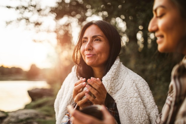 older woman with long dark hair outside with sunsetting behind her and smiling holding a cup
